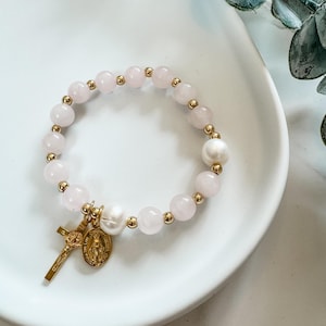 May include: A bracelet with light pink rose quartz beads, gold-coloured spacers, and a gold cross charm with a religious medallion. It also features a large white pearl.