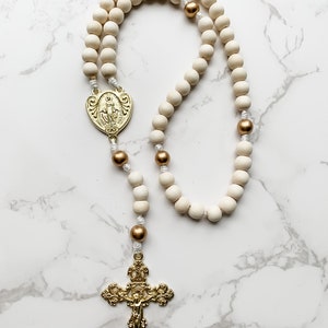 The wood bead rosary lays flat and the entire rosary is visible. The crucifix and centerpiece are shiny and gold and the wood beads are white and gold.