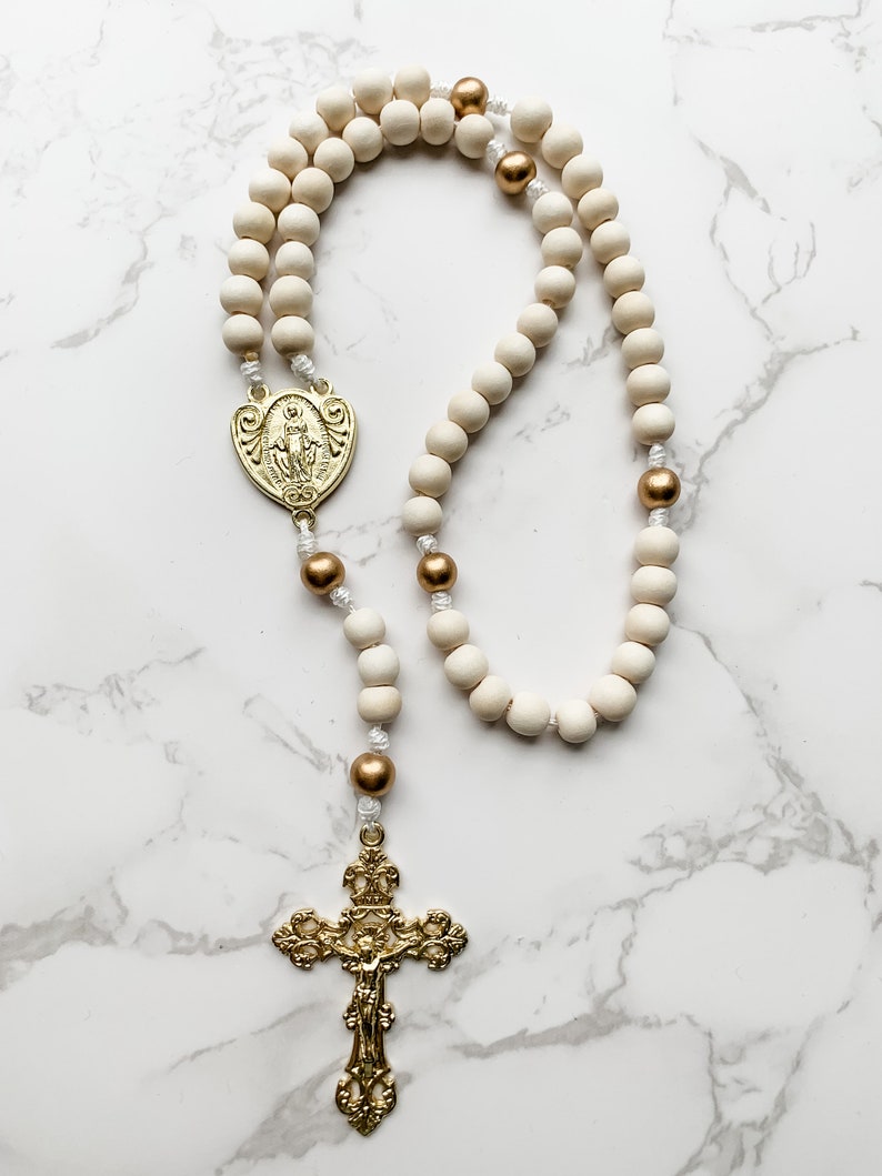 The wood bead rosary lays flat and the entire rosary is visible. The crucifix and centerpiece are shiny and gold and the wood beads are white and gold.