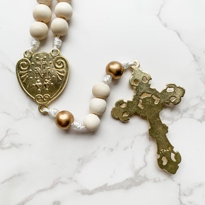 Close up image of the back of the crucifix and centerpiece of the rosary. The back of the gold crucifix is flat and the word Italy is visible near the bottom. The back of the Miraculous Medal depicts two hearts.
