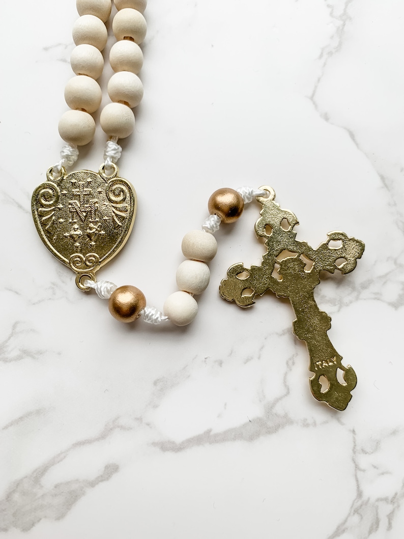 Close up image of the back of the crucifix and centerpiece of the rosary. The back of the gold crucifix is flat and the word Italy is visible near the bottom. The back of the Miraculous Medal depicts two hearts.