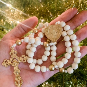 The white and gold rosary lays in the palm of a woman’s hand. A Christmas tree and white lights are visible in the background.