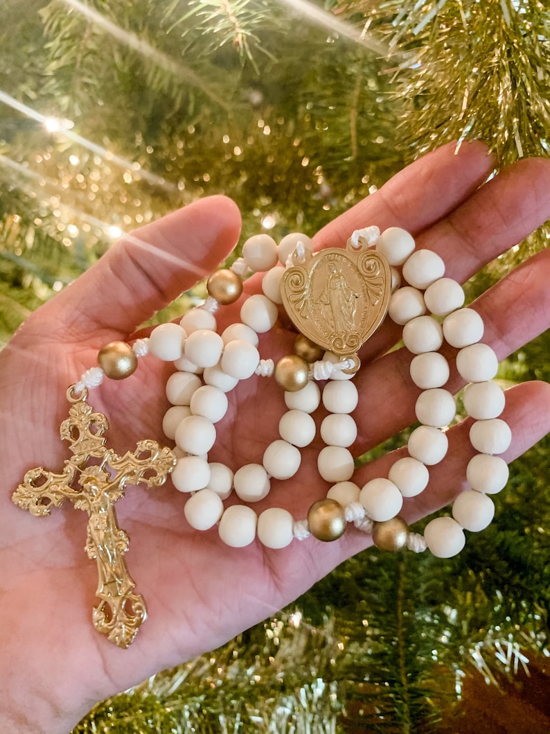 The white and gold rosary lays in the palm of a woman’s hand. A Christmas tree and white lights are visible in the background.