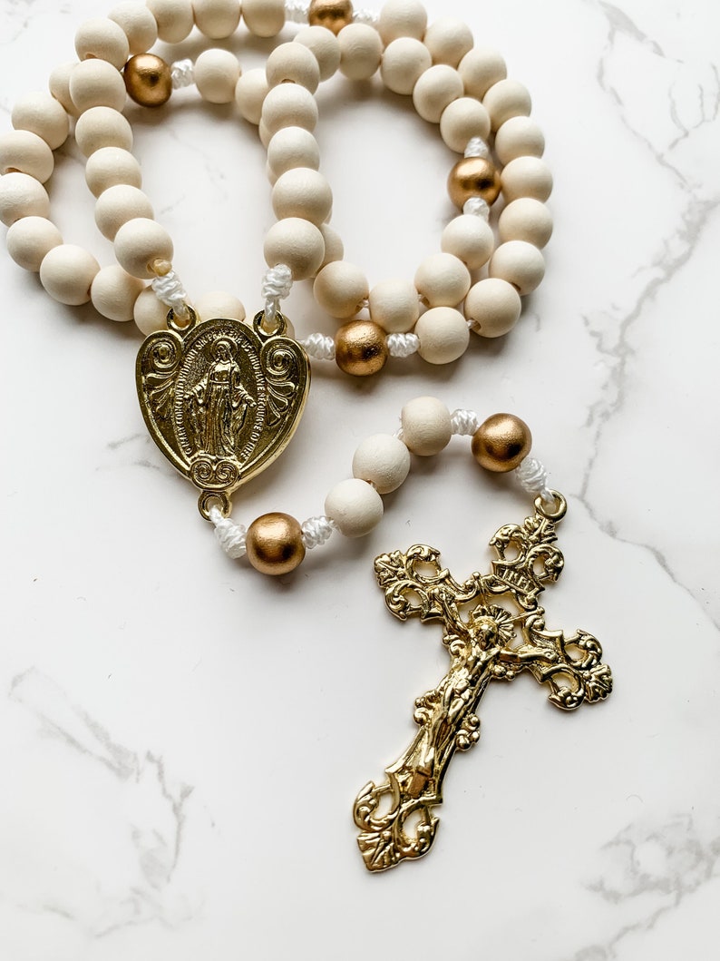 Close up image of a wood bead rosary. The crucifix is shiny and gold with an elaborate fleur de lis design. The beads of the rosary are white and gold. The centerpiece is a slightly heart shaped Miraculous Medal.