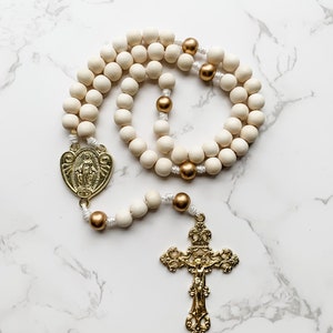 The wood bead rosary lays in a curled up arrangement and the entire rosary is visible. The crucifix and centerpiece are shiny and gold and the wood beads are white and gold.