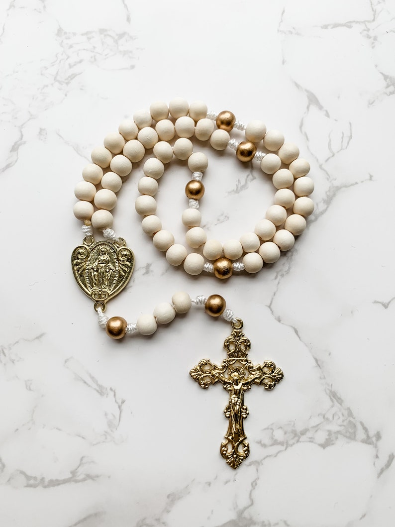 The wood bead rosary lays in a curled up arrangement and the entire rosary is visible. The crucifix and centerpiece are shiny and gold and the wood beads are white and gold.