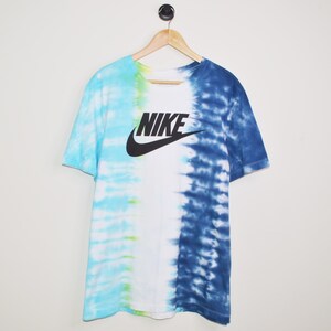 May include: A white t-shirt with a blue and light blue tie-dye pattern on the right side and a light blue and green tie-dye pattern on the left side. The shirt has a black Nike logo with the word "NIKE" in black letters.