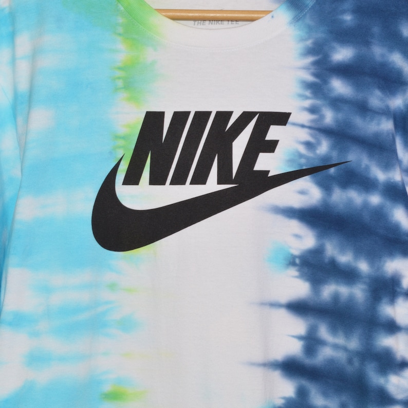 May include: A white t-shirt with a tie-dye pattern in blue and green. The Nike logo is printed in black on the front of the shirt.