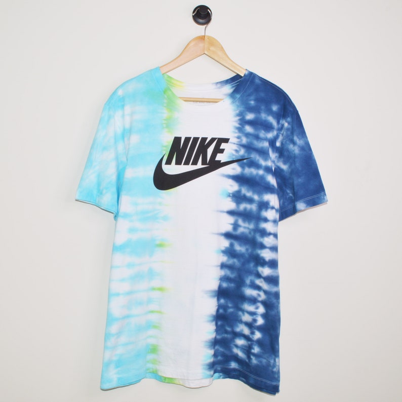 May include: A white t-shirt with a blue and light blue tie-dye pattern on the right side and a light blue and green tie-dye pattern on the left side. The shirt has a black Nike logo with the word "NIKE" in black letters.