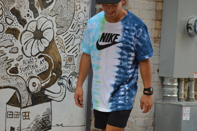 May include: A person wearing a blue and white tie-dye t-shirt with a black Nike logo and the word "NIKE" in white. The person is wearing black shorts and a black watch.