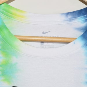 May include: A white t-shirt with a tie-dye pattern in shades of blue, green, and yellow. The shirt has a Nike logo on the front and the text "Nike Tee" and "Made in Nicaragua" on the tag.