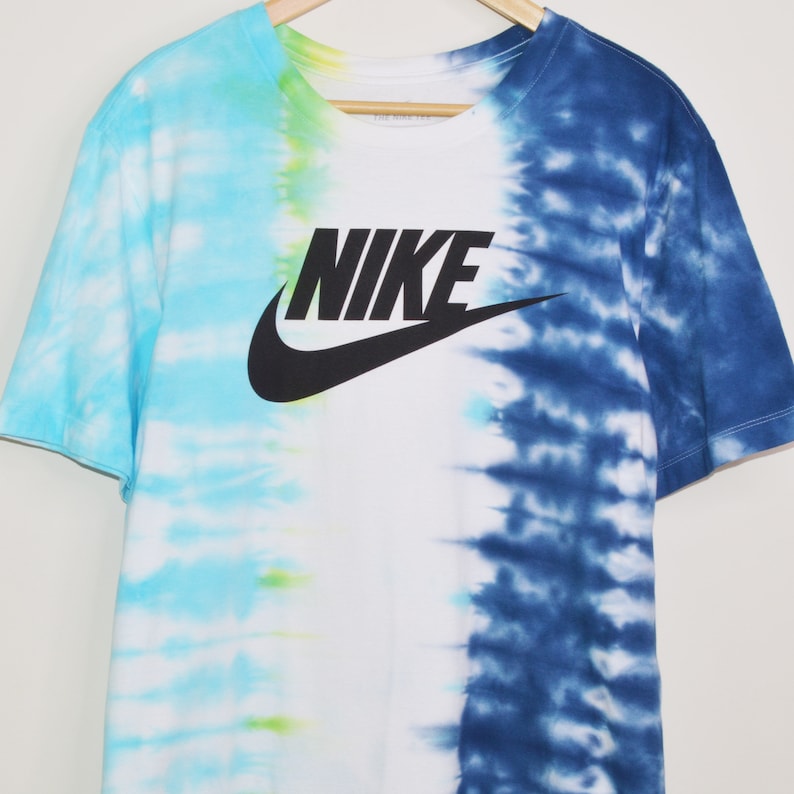 May include: A white t-shirt with a blue and light blue tie-dye pattern on the right side and a light blue and green tie-dye pattern on the left side. The shirt has a black Nike logo with the word "NIKE" in white letters.