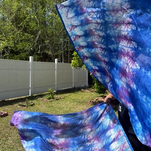 May include: Two flowing fabric flags with a tie-dye pattern in shades of blue and purple are held outdoors. The flags are billowing in the wind against a backdrop of a white fence, green grass, and trees.