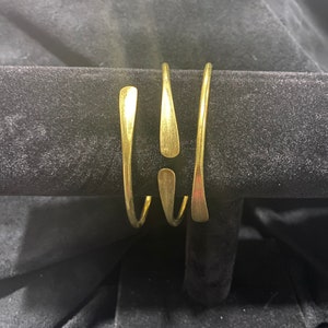 May include: A gold-colored cuff bracelet with a unique design. The bracelet features three curved, elongated shapes that wrap around the wrist. The metal has a polished finish, reflecting light. The bracelet is displayed on a black velvet surface.