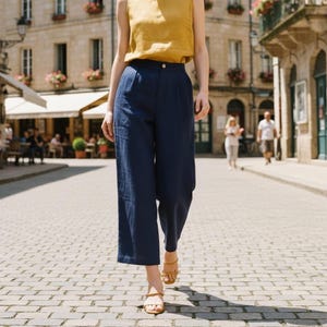 May include: A woman in a mustard yellow sleeveless top and navy blue wide-leg trousers walks along a cobbled street. She wears tan sandals. The background shows a European-style building with flower boxes and a cafe.