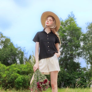 May include: A woman wearing a black short-sleeved button-down shirt, white shorts, brown boots, and a straw hat with a brown scarf tied around her head. She is holding a bouquet of red flowers in her right hand. The woman is standing in a field of dry grass.