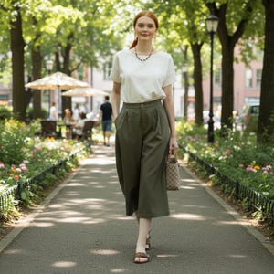 May include: A person in a white top and olive green culottes strolls along a path in a park. They carry a patterned handbag and wear brown sandals. A necklace and earrings are visible. The background features trees, flowers, and other people enjoying the outdoors.