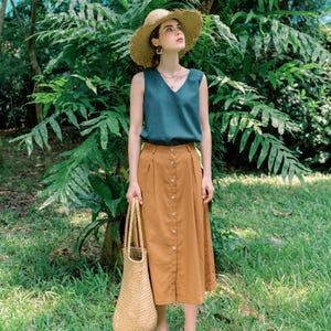 May include: A woman wearing a teal sleeveless top and a brown button-down skirt. She is holding a woven straw bag and wearing a wide-brimmed straw hat.