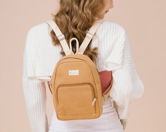 May include: A tan canvas backpack with a white strap and a small white label that says "The Daily Edited".