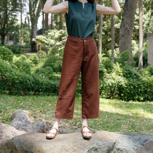 May include: A woman wearing a green sleeveless top, brown wide-leg trousers, and a straw hat. She is standing on a large rock in a park setting.