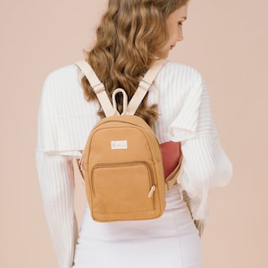 May include: A tan canvas backpack with a white strap and a small white label that says "The Daily Edited".