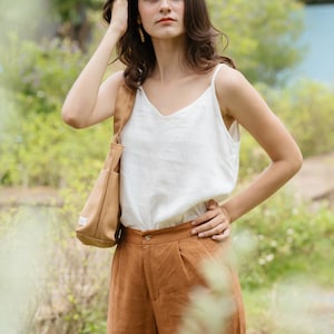 May include: A woman wearing a white tank top and brown pants with a brown tote bag slung over her shoulder. She is standing in a grassy area with green foliage in the background.