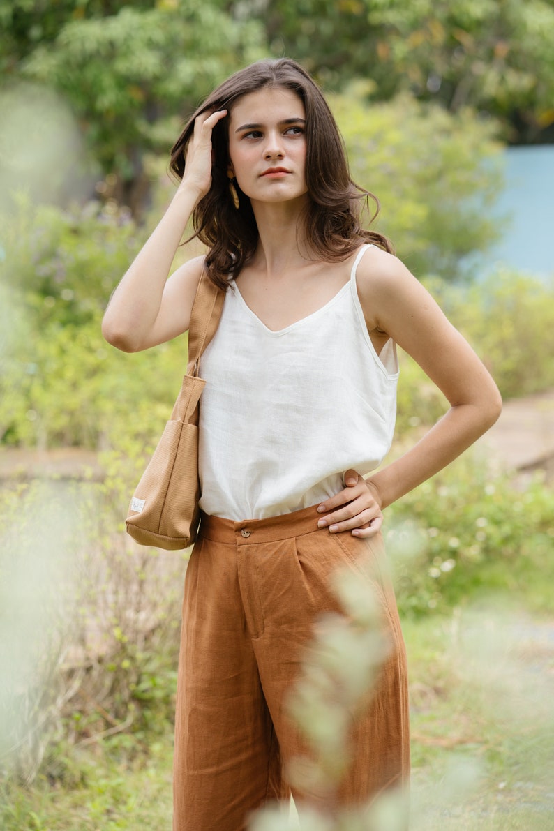 May include: A woman wearing a white tank top and brown pants with a brown tote bag slung over her shoulder. She is standing in a grassy area with green foliage in the background.