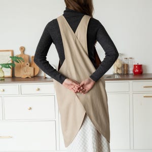May include: A beige cross-back apron with a unique design. The apron is made of a light-colored fabric and is worn over a dark gray long-sleeved shirt. The apron's straps cross in the back. The person is standing in a kitchen setting.