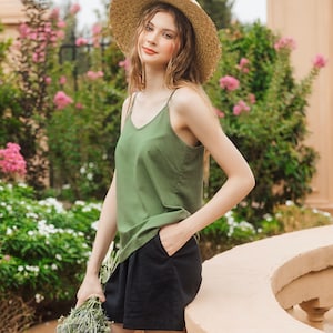 Op de afbeelding: Een vrouw in een groene tanktop en een zwarte broek, met een brede strohoed, staat in een tuin.
