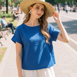 May include: A woman wearing a straw hat and a blue short-sleeved top with white shorts. The hat has a wide brim. The top is a solid blue color. The shorts are white. The background is a city street with trees and buildings.