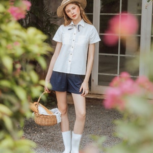 May include: A woman wearing a white button-down shirt with a blue and white scarf tied around the collar, blue denim shorts, and white rain boots. She is holding a wicker basket with a loaf of bread inside. The woman is standing in front of a brick wall with a window behind her.