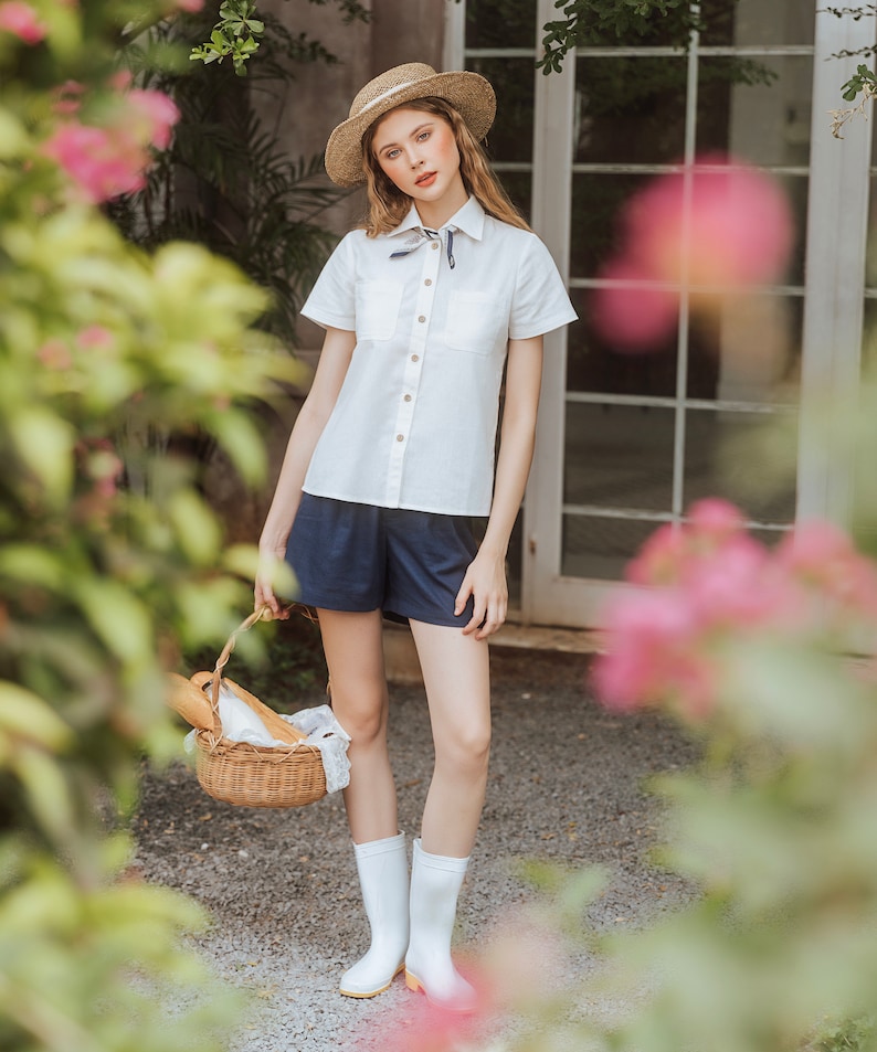 May include: A woman wearing a white button-down shirt with a blue and white scarf tied around the collar, blue denim shorts, and white rain boots. She is holding a wicker basket with a loaf of bread inside. The woman is standing in front of a brick wall with a window behind her.