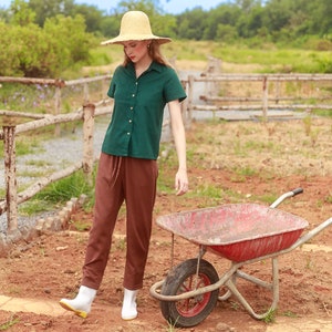 May include: A woman wearing a green button-down shirt, brown pants, and white boots stands in a field with a red wheelbarrow. She is wearing a straw hat and looking down.