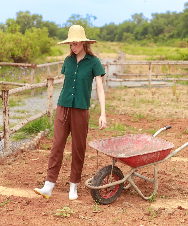 May include: A woman wearing a green button-down shirt, brown pants, and white boots stands in a field with a red wheelbarrow. She is wearing a straw hat and looking down.