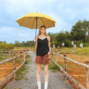 Op de afbeelding: Een vrouw in een zwart tanktop en een bruine short staat op een grindpad en houdt een gele paraplu vast. Ze draagt witte laarzen en heeft lang blond haar. Het pad is omzoomd met houten hekken en er zijn bomen en struiken op de achtergrond.