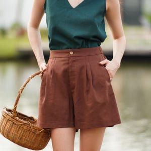 May include: Brown linen shorts with pleats and a button fastening. The shorts are worn with a dark green sleeveless top.