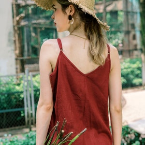 Op de afbeelding: Een vrouw draagt een rood tanktop, een beige broek en een strohoed met een brede rand. Ze houdt een bos groene takken vast.