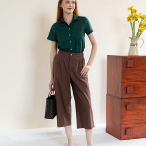 May include: A woman in a dark green short-sleeved shirt and brown culottes, holding a black woven tote bag and wearing black sandals. A wooden chest of drawers and a vase of yellow flowers are in the background.