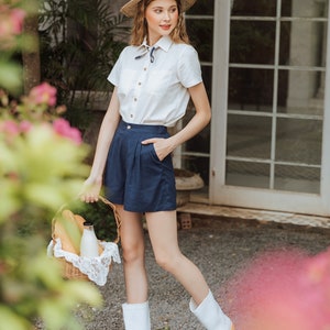 May include: A woman wearing a white button-down shirt with a blue and white scarf, blue shorts, and white rain boots. She is holding a wicker basket with bread and a bottle of milk.