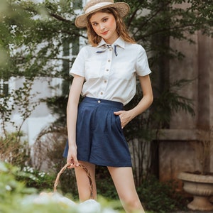 May include: A woman wearing a white button-down shirt with a blue and white patterned scarf, blue linen shorts, and a straw hat. She is holding a wicker basket.
