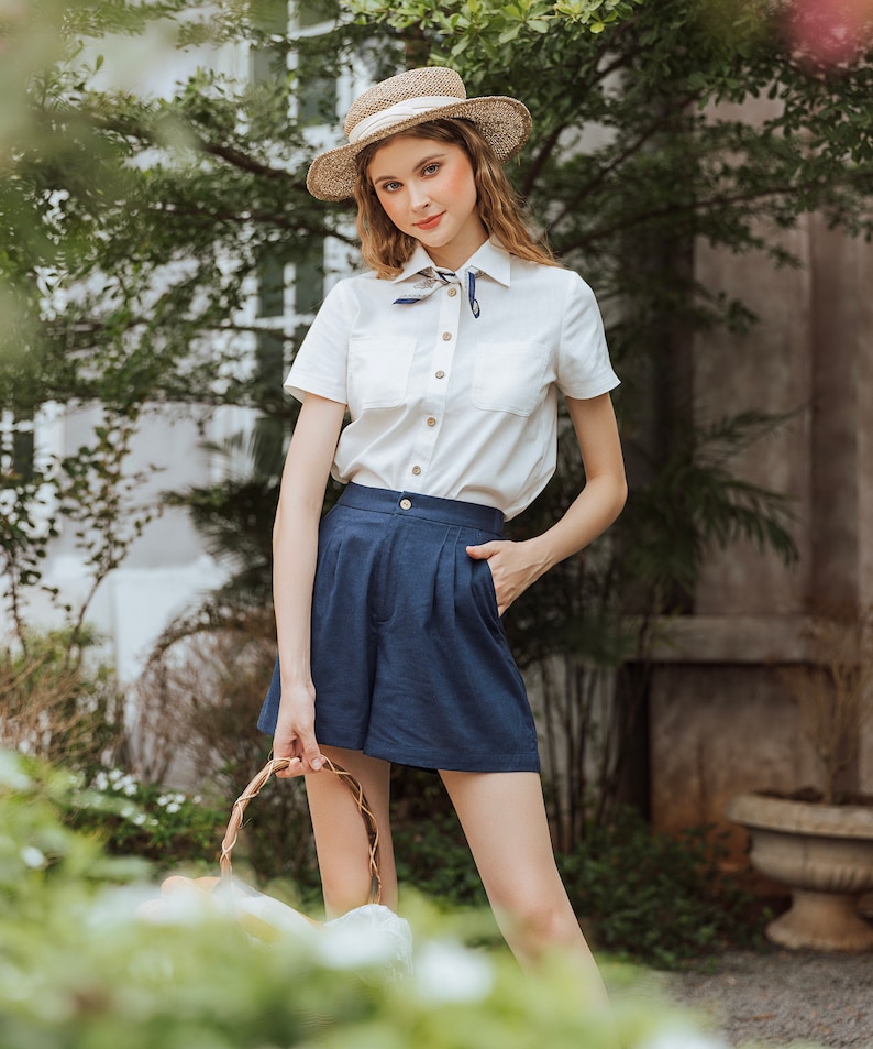 May include: A woman wearing a white button-down shirt with a blue and white patterned scarf, blue linen shorts, and a straw hat. She is holding a wicker basket.