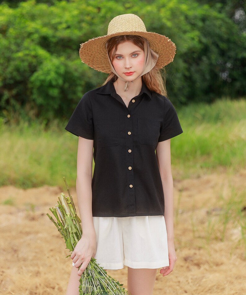 May include: A woman wearing a black collared shirt with short sleeves and white shorts. She is wearing a straw hat with a light brown ribbon. She is holding a bouquet of green flowers.