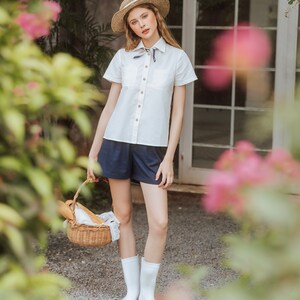 May include: A woman wearing a white button-down shirt with a blue and white scarf tied around the collar, blue denim shorts, and white rain boots. She is holding a wicker basket with a loaf of bread inside. The woman is standing in front of a brick wall with a window behind her.