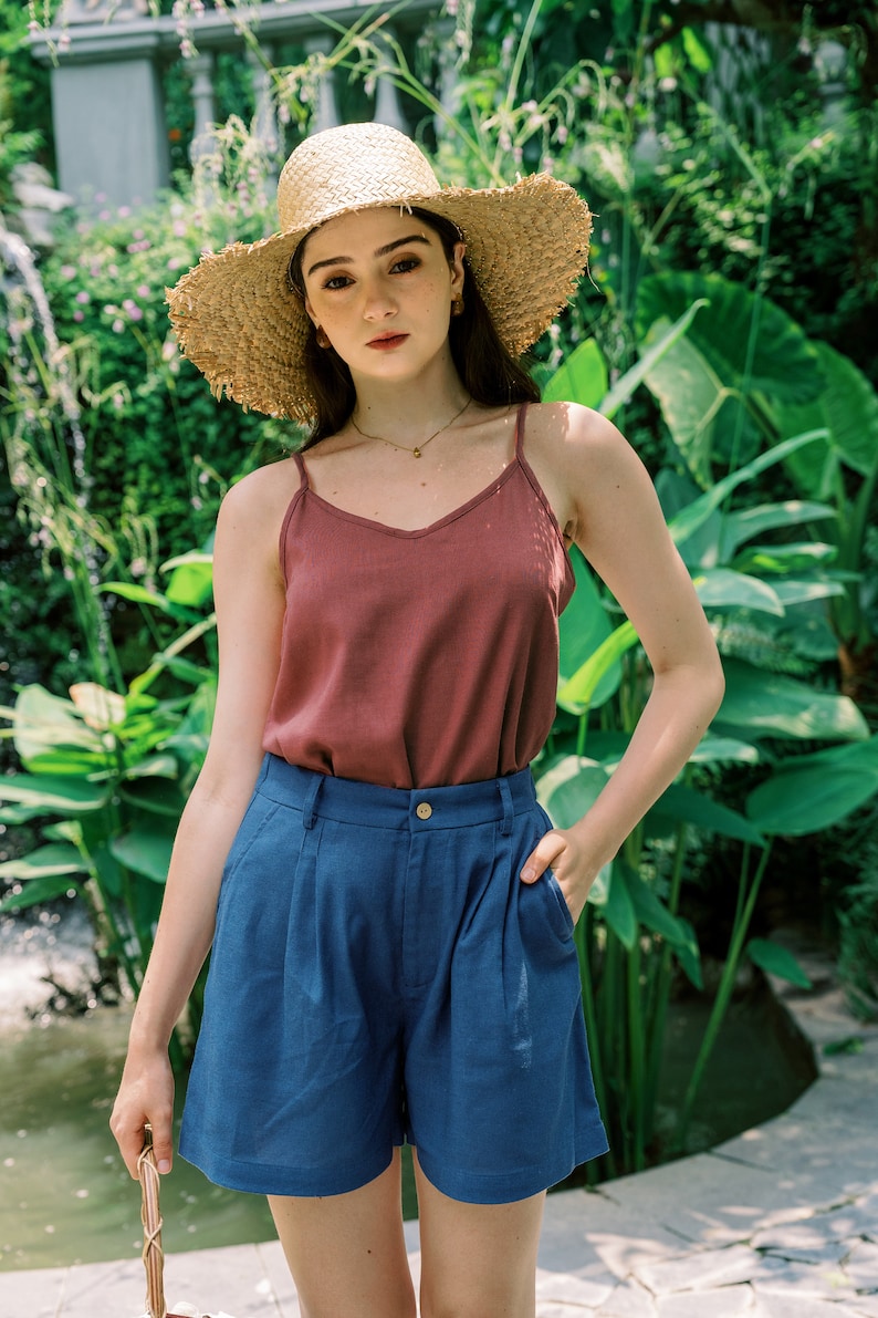 Op de afbeelding: Een vrouw draagt een strohoed, een bordeauxrode tanktop en een blauwe denim short. Ze staat voor een weelderige groene achtergrond.