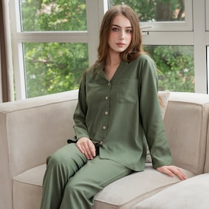 May include: Olive green pyjama set. The set features a long-sleeved button-down shirt with a chest pocket and matching trousers. The pyjamas are made of a soft, comfortable fabric. The model is sitting on a beige corduroy sofa.