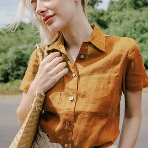 May include: A woman wearing a brown linen shirt with a patch pocket and short sleeves. She is holding a woven straw bag.
