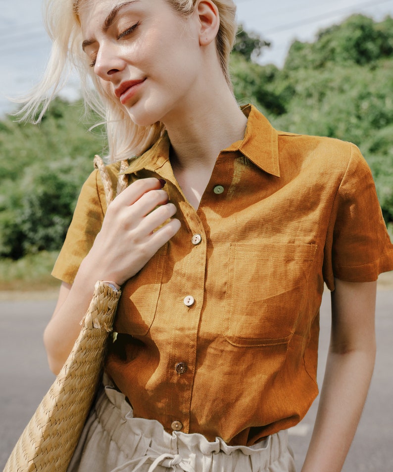 May include: A woman wearing a brown linen shirt with a patch pocket and short sleeves. She is holding a woven straw bag.