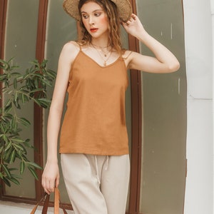 May include: A woman wearing a brown sleeveless top with a v-neck and a straw hat. She is wearing beige pants and holding a woven tote bag.