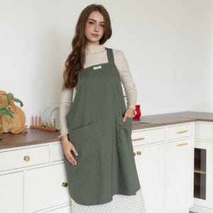 May include: An olive green apron with large front pockets and a square neckline. The apron is worn over a cream-colored long-sleeved shirt. The apron has a small label on the chest. The apron is made of a textured fabric.