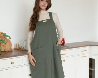 May include: An olive green apron with large front pockets and a square neckline. The apron is worn over a cream-colored long-sleeved shirt. The apron has a small label on the chest. The apron is made of a textured fabric.